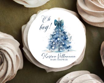 May include: A round white sticker with a blue Christmas tree design, a blue bow, and the words "Oh boy!" in script. The sticker also includes the text "Korina Villanueva BABY SHOWER". The sticker is surrounded by white, swirled objects.