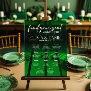 Puede incluir: Un plano de asientos de boda de acrílico verde sobre un caballete de madera. El letrero dice "find your seat and bon appetit" sobre los nombres Olivia & Daniel y la fecha 26 de agosto de 2028. Los nombres de los invitados se enumeran debajo de los números de mesa.