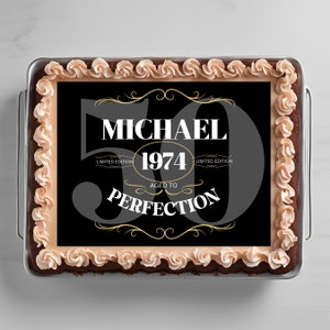 Può includere: Una torta quadrata nera e dorata con uno sfondo nero e testo bianco che recita "Michael 1974 Aged to Perfection" con le parole "Edizione limitata" su entrambi i lati dell'anno. La torta è decorata con un bordo di glassa marrone e un design a spirale di glassa beige.