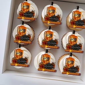 May include: A box of nine white cupcakes decorated with edible images of a bottle of liquor, a glass of liquor, and a cigar. The images are all in gold, black, and brown tones. The cupcakes are decorated with the year "1979" and the name "Jonathan".