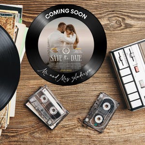 May include: A black vinyl record with a photo of a couple, text that reads "COMING SOON" and "SAVE the DATE", and the names "Mr. and Mrs. Anderson". The record is on a wooden surface with cassette tapes and a cassette player.