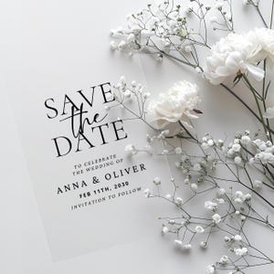 Puede incluir: Una tarjeta transparente de Save the Date con texto negro, con las palabras "SAVE the DATE" y detalles de la boda. Flores blancas y vegetación se disponen alrededor de la tarjeta, creando una estética suave y elegante.