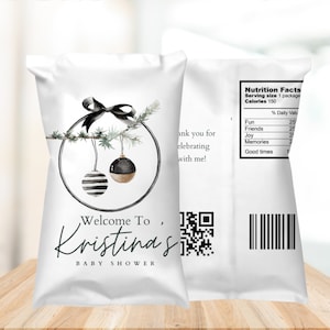 May include: White favor bag with a black and white striped ornament and a gold ornament hanging from a green pine branch. The bag has a black ribbon and the text "Welcome To, Kristina's Baby Shower".