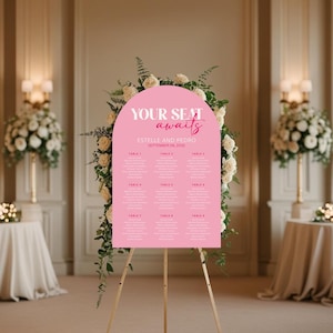 May include: A pink wedding seating chart with the words "YOUR SEAT awaits" and the names "ESTELLE AND PEDRO" and the date "SEPTEMBER 08, 2020". The chart is on a wooden easel, surrounded by white flowers.