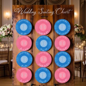 Op de afbeelding: Een houten trouwzittingkaart met de tekst "Wedding Seating Chart" in elegant schrift. De kaart bevat blauwe en roze vinylplaatvormige placeholders met gastnamen en tafelopdrachten. De platen hebben zinnen als "Just the two of us".