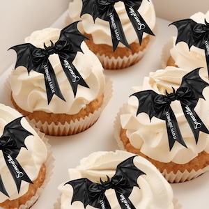 Op de afbeelding: Zes cupcakes versierd met zwarte vleermuisvormige cupcaketoppers met zwarte strikken en de tekst "Happy Birthday Samantha".
