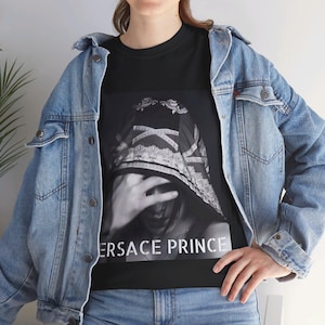May include: Black t-shirt with a graphic print of a person wearing a patterned head covering. The text "VERSACE PRINCE" is printed on the bottom of the shirt.