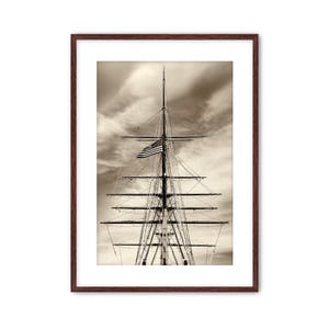 May include: Sepia-toned framed print of a ship's mast with an American flag waving in the wind. The mast is tall and detailed, with multiple horizontal spars and rigging. The sky is cloudy, adding depth to the nautical scene.