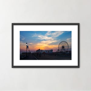 May include: A framed photograph of a boardwalk at sunset. The sky is a mix of orange, yellow, and blue. A Ferris wheel is visible in the distance, along with other buildings and people.