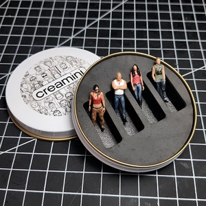 May include: A white round tin with a black and white illustration of faces and the text "Creamini" on the lid. The tin is open and reveals a black foam insert with four empty slots for figurines.