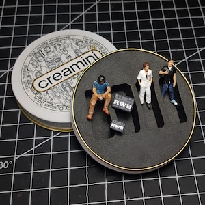 May include: A round tin container with a black foam interior. The tin has a white label with the text "creamini" and "high quality miniatures hand painted". The foam interior has three miniature figures and two black cubes with the text "RWB" on them.