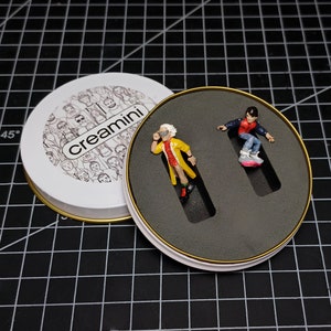 May include: A white, round tin with a gold rim and the word "creamini" on the lid. Inside, a black insert holds two miniature figurines: one in a yellow coat and the other on a hoverboard. The background is a black grid.