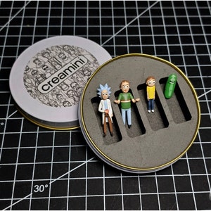 Puede incluir: Una lata redonda blanca con las palabras "Creamini" impresas en ella. La lata está abierta y muestra un inserto de espuma negra con cinco pequeñas figuritas. Las figuritas son un pepinillo, un personaje de dibujos animados con cabello amarillo, un personaje de dibujos animados con cabello verde, un personaje de dibujos animados con cabello marrón y un personaje de dibujos animados con cabello azul.