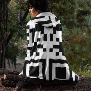 Puede incluir: Una persona lleva una manta con capucha negra y blanca con un patrón de código QR. La manta está hecha de vellón suave y tiene una capucha cómoda. La persona está sentada en un tronco en un entorno boscoso.