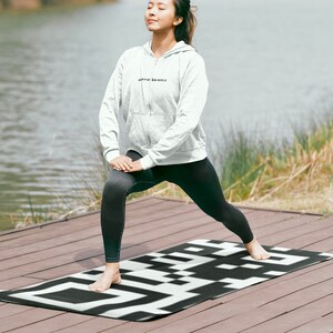 May include: A person in a white hooded top and black leggings practices yoga on a black and white geometric patterned mat. The mat is on a wooden deck near a lake. The top has text on it.
