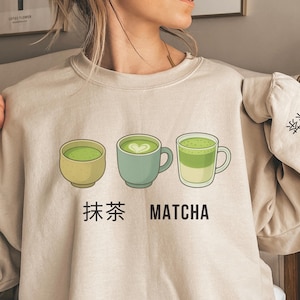 May include: Beige sweatshirt with three matcha tea cups in green and yellow, with the Japanese characters for matcha and the English word "Matcha" printed below. A matcha cup illustration is on the sleeve.