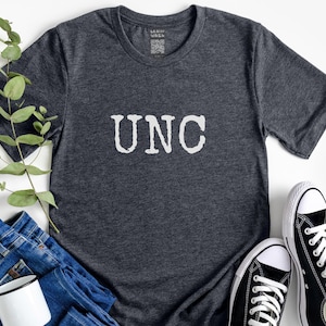May include: Dark gray t-shirt with the letters "UNC" in white, paired with blue jeans, a white mug, and black and white sneakers. A sprig of greenery adds a natural touch to the casual arrangement.