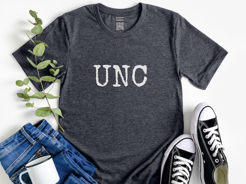 May include: Dark gray t-shirt with the letters "UNC" in white, paired with blue jeans, a white mug, and black and white sneakers. A sprig of greenery adds a natural touch to the casual arrangement.