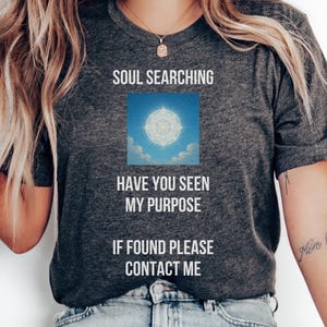 Puede incluir: Una camiseta gris oscuro con el texto "Soul Searching" y un gráfico de un mandala en un cielo azul. El texto debajo del gráfico dice "¿Has visto mi propósito? Si lo encuentras, por favor contacta conmigo."