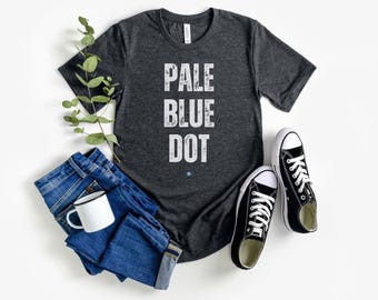Puede incluir: Camiseta gris oscuro con el texto blanco "PALE BLUE DOT", combinada con vaqueros azules, zapatillas negras y una taza blanca. Una ramita de vegetación añade un toque natural al conjunto informal.
