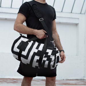 Könnte beinhalten: Eine schwarz-weiße geometrisch gemusterte Reisetasche mit einem schwarzen Schultergurt. Die Tasche wird von einer Person getragen, die ein schwarzes T-Shirt und schwarze Shorts trägt.