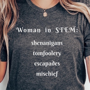 May include: Dark gray t-shirt with the text "Woman in STEM: shenanigans, tomfoolery, escapades, mischief" in white. The shirt has a crew neck and short sleeves. A gold necklace is visible.