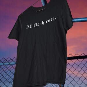 K&ouml;nnte beinhalten: Schwarzes T-Shirt mit dem Schriftzug "All flesh rots" in wei&szlig;er gotischer Schrift. Das Shirt h&auml;ngt an einem Metallb&uuml;gel vor einem Hintergrund aus Maschendrahtzaun und Sonnenuntergang.
