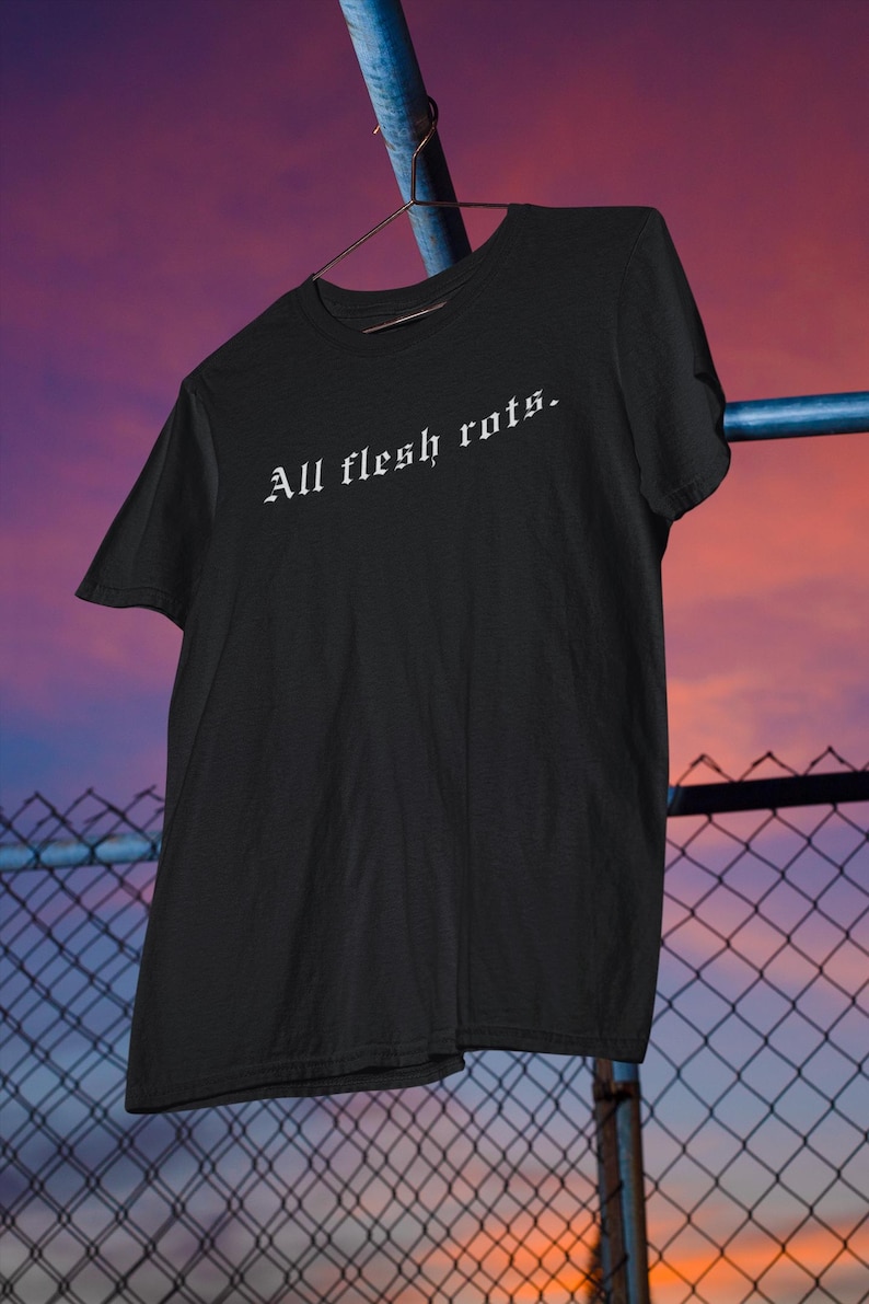 K&ouml;nnte beinhalten: Schwarzes T-Shirt mit dem Schriftzug "All flesh rots" in wei&szlig;er gotischer Schrift. Das Shirt h&auml;ngt an einem Metallb&uuml;gel vor einem Hintergrund aus Maschendrahtzaun und Sonnenuntergang.