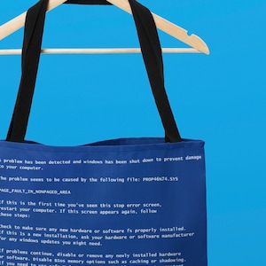 Op de afbeelding: Een blauwe tote bag met zwarte banden en een witte handgreep. De tas heeft een blauw scherm dat op de voorkant is bedrukt met de tekst "Er is een probleem gedetecteerd en Windows is afgesloten om schade aan uw computer te voorkomen." De tekst gaat verder met "Het probleem lijkt te worden veroorzaakt door het volgende bestand: PROP46N74.SYS PAGE_FAULT_IN_NONPAGED_AREA".