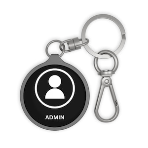 May include: A silver keychain with a black round tag that says "ADMIN" and has a white silhouette of a person. The keychain has a silver ring and a silver clip.