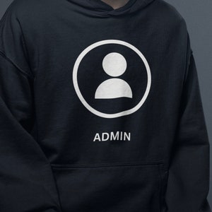 Peut inclure: Un sweat à capuche noir avec un motif blanc représentant une personne dans un cercle, avec le mot "ADMIN" en dessous. Le sweat à capuche a une poche avant et une capuche. Le motif est centré.