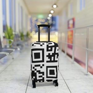 May include: A black and white suitcase cover with a repeating QR code pattern. The suitcase is standing on a tiled floor in an airport terminal.