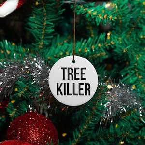 Op de afbeelding: Ronde witte kerstbal met zwarte tekst "TREE KILLER" die aan een groene kerstboom hangt.