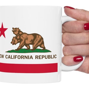 May include: White ceramic mug with a red star, a brown bear, and the text "NEW CALIFORNIA REPUBLIC" printed on the front. The mug is being held by a hand with red nail polish.