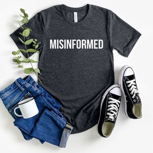 May include: A dark gray t-shirt with the word "MISINFORMED" printed in white on the front. The shirt is laid flat on a white surface with a pair of black canvas shoes, a pair of blue jeans, and a sprig of greenery.