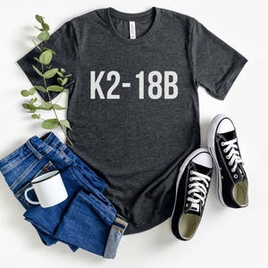 May include: A dark heather grey t-shirt with the text "K2-18B" printed in white on the front. The shirt is paired with blue jeans and black sneakers.