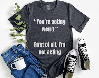 Puede incluir: Camiseta gris oscuro con el texto "You're acting weird. First of all, I'm not acting." La imagen incluye vaqueros azules, una taza blanca, una ramita verde y zapatillas deportivas blancas y negras.