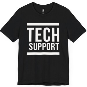 以下が含まれることがあります： 黒いTシャツ。白い文字で「TECH SUPPORT」と大きく書かれています。テキストは中央に配置され、水平の白い線で囲まれています。Tシャツはクルーネックで半袖です。