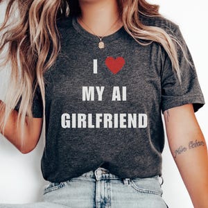 以下が含まれることがあります： 白い文字で「I ♥ MY AI GIRLFRIEND」とプリントされた濃いグレーのTシャツ。ハートは赤色です。