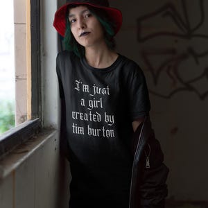 May include: A person wearing a black t-shirt with the text "I'm just a girl created by Tim Burton" in white gothic font. They are wearing a red and black bucket hat and a black leather jacket.
