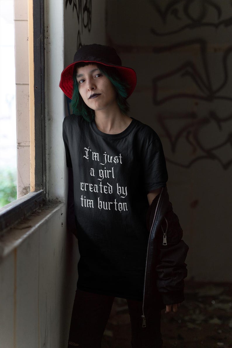 May include: A person wearing a black t-shirt with the text "I'm just a girl created by Tim Burton" in white gothic font. They are wearing a red and black bucket hat and a black leather jacket.