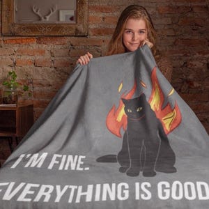 Puede incluir: Una manta gris con una ilustración de un gato negro rodeado de llamas. El texto blanco dice "I'M FINE. EVERYTHING IS GOOD.". Una persona sostiene la manta.