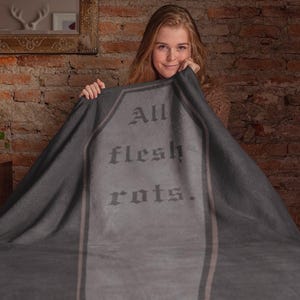May include: Dark gray blanket with the text "All flesh rots" in a gothic font. The blanket has a tombstone design with vertical stripes. The blanket is held up by a person.