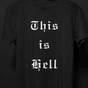 May include: Dark gray t-shirt with the words "This is Hell" printed in white gothic font. The text is stacked vertically, with each word centered on the shirt. The shirt has a crew neck and short sleeves.