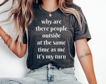 Może przedstawiać: Ciemnoszara koszulka z białym napisem "why are there people outside at the same time as me it's my turn". Koszulka jest noszona z niebieskimi dżinsami.