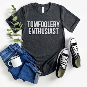 Op de afbeelding: Donkergrijs t-shirt met de witte tekst "TOMFOOLERY ENTHUSIAST", gecombineerd met een blauwe jeans, een witte mok en zwarte canvas sneakers. Een casual outfit.