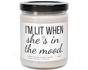 May include: A white glass candle jar with a black lid and a white label that reads "I'M LIT WHEN she's in the mood." The label also includes the text "100% NATURAL VEGAN SOY WAX - ECO-FRIENDLY - NON-TOXIC - REUSABLE GLASS JAR - BE THE SPARK"