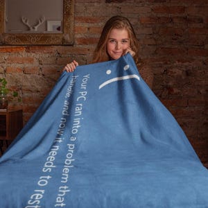 Könnte beinhalten: Eine blaue Decke mit dem Text "Your PC ran into a problem that handle, and now it needs to rest" und einem traurigen Gesicht. Eine Person hält die Decke.