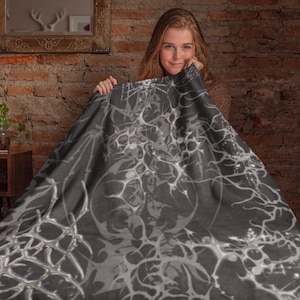 May include: A dark gray blanket with a white, abstract design. The blanket is draped, revealing a portion of the design. The background includes a brick wall, a wooden table, and a mirror.