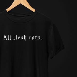 K&ouml;nnte beinhalten: Schwarzes T-Shirt mit dem gotischen Text "All flesh rots." Das Shirt h&auml;ngt an einem Holzb&uuml;gel vor einem dunklen Hintergrund. Der Text ist in einer fetten, wei&szlig;en Schrift.